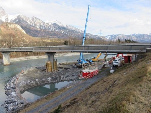Baustelle Kolksicherung an der Rheinbrücke Fläsch: Pfeiler mit Spundwandumschliessung im Rhein, Blocksteinschüttung am Ufer, Autokran und Bagger unter der Brücke, verschneite Berge im Hintergrund
